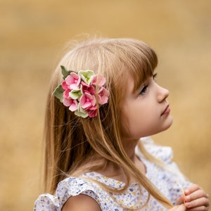 May include: A young girl with long blonde hair wearing a floral dress and a pink and green felt flower hair clip.