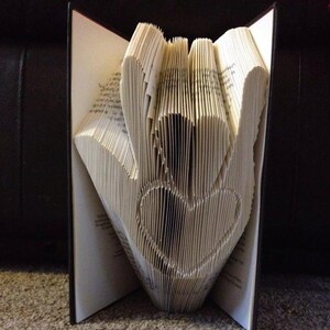Könnte beinhalten: Ein offenes Buch, dessen Seiten zu einer Hand mit einem Herz gefaltet sind. Die Falttechnik ist deutlich zu erkennen. Die Seiten sind hellbeige, der Buchrücken schwarz. Das Buch liegt auf einem Teppich.