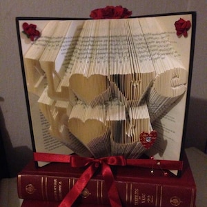 Puede incluir: Una escultura de arte de libro de la palabra "LOVE" hecha de páginas dobladas de un libro. La escultura está decorada con rosas rojas y una cinta roja. La escultura está sentada encima de una pila de libros rojos.