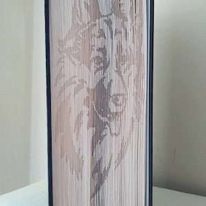 May include: A book with pages folded and cut to create a 3D image of a wolf's face. The book is bound in a dark blue cover.