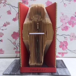 May include: A book sculpture of a person standing with a red and gold book cover. The sculpture is on a gray surface with the text "David Q.S. ?" on the bottom.