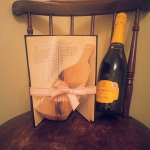 May include: A book folded into the shape of a cat, decorated with a white ribbon with red stitching, is displayed on a wooden chair with a bottle of prosecco.