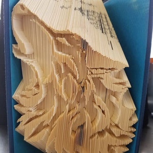 May include: An open book with pages intricately folded to create a three-dimensional sculpture. The pages are a light beige color, and the book's cover is blue. The folded pages form a detailed, abstract design.