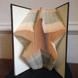 Puede incluir: Una escultura de arte de libro en forma de estrella de mar hecha doblando las páginas de un libro. La escultura está hecha de un libro con una cubierta negra y las páginas son de color crema.