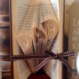 May include: A book art sculpture, also known as a book folding, in the shape of a heart. The book is bound in brown leather and has a brown ribbon tied around it. The book is sitting on a wooden surface.