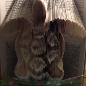 May include: A book folded into the shape of a turtle. The turtle is made of paper and is a light brown colour. The book is decorated with a green ribbon with white and pink polka dots.