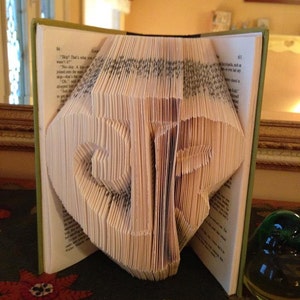 Könnte beinhalten: Ein Buch mit Seiten, die zu einer dreidimensionalen Skulptur des Wortes "ZIP" gefaltet sind. Das Buch ist geöffnet und die Skulptur ist von vorne sichtbar. Das Buch liegt auf einer grün-braunen gemusterten Oberfläche.