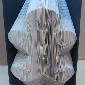 May include: A book folded into the shape of a gingerbread person, standing on a black base with a gingerbread person ribbon.
