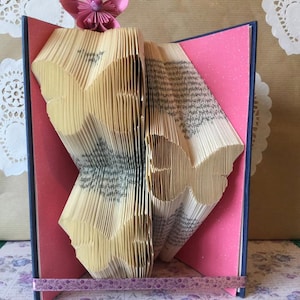 May include: A book with pages folded into the shape of a butterfly, decorated with a pink origami flower on top. The book is open and the pages are folded into a 3D butterfly shape. The book is sitting on a pink and white patterned surface.
