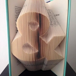 May include: A book with pages folded into the shape of a letter 'B'. The book is open and the pages are folded to create a three-dimensional shape. The book is on a black surface and the cover is a light blue color. The text on the cover of the book reads "BANK".