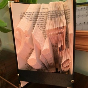 May include: A book art sculpture, also known as a book folding, in the shape of the letter "S". The book is bound in black with gold accents and a black ribbon. The pages are folded to create a three-dimensional shape.
