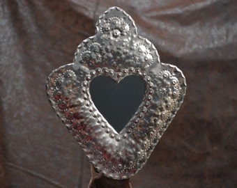 May include: A silver heart-shaped mirror with a decorative, embossed design. The mirror is surrounded by a frame with a scalloped edge.