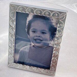 May include: A silver picture frame with a decorative embossed border. The frame holds a black and white photo of a young child smiling.