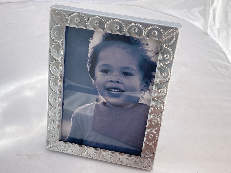 May include: A silver picture frame with a decorative embossed border. The frame holds a black and white photo of a young child smiling.
