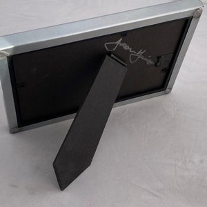 May include: A silver metal picture frame with a black mat and a black easel stand. The frame has a signature in silver ink that reads "Jenny" on the front.