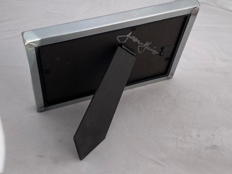 May include: A silver metal picture frame with a black mat and a black easel stand. The frame has a signature in silver ink that reads "Jenny" on the front.