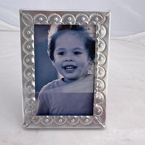 May include: Silver picture frame with a decorative embossed border. The frame holds a black and white photo of a young child smiling.