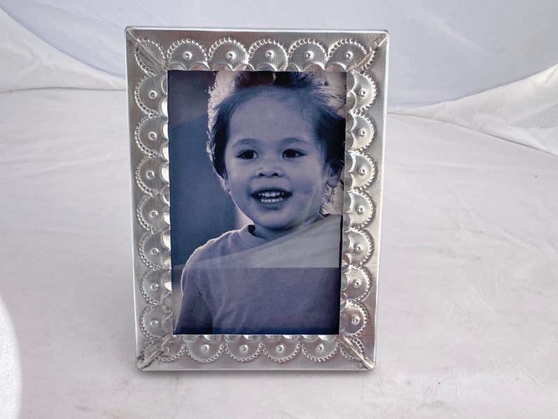 May include: Silver picture frame with a decorative embossed border. The frame holds a black and white photo of a young child smiling.