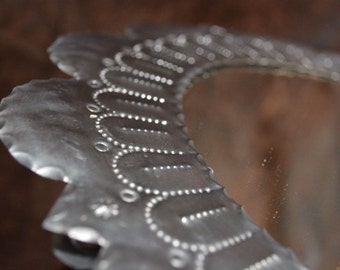 May include: A silver metal mirror with a decorative scalloped edge. The mirror has a floral design with small circles around the edge.