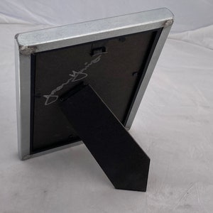 May include: A silver picture frame with a black mat and a black easel stand. The frame is empty and has a signature in the mat.