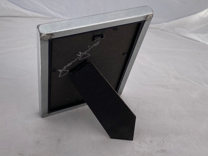 May include: A silver picture frame with a black mat and a black easel stand. The frame is empty and has a signature in the mat.