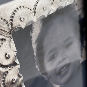 May include: A silver metal picture frame with a decorative embossed design. The frame holds a black and white photo of a young child smiling.