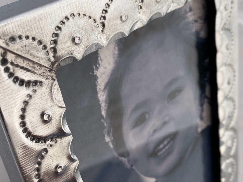 May include: A silver metal picture frame with a decorative embossed design. The frame holds a black and white photo of a young child smiling.