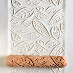 May include: A wooden rolling pin with a leaf pattern carved into the surface. The rolling pin is resting on a white surface with a leaf pattern embossed into it.