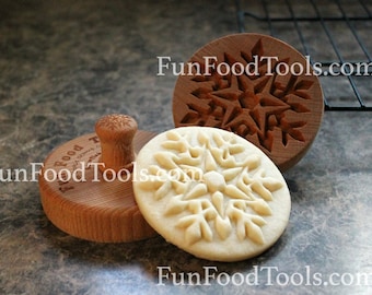 May include: A wooden cookie press with a snowflake design, alongside a pressed cookie and the matching mold. The press has a round handle with a snowflake carving. The text "FunFoodTools.com" is visible.