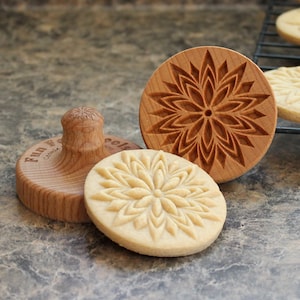 Könnte beinhalten: Zwei Holz-Keksstempel mit einem floralen Design und ein Keks mit dem gleichen Design. Die Keksstempel sind aus Holz und haben eine natürliche Oberfläche. Der Keks ist hellbraun und hat eine leicht knusprige Textur.