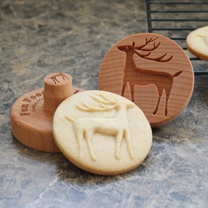 May include: Wooden cookie stamps and stamped cookies with a deer design. One stamp is round with a deer carving, another is a handle with a deer, and the third is a round stamp with the text "Fun Food". The cookies are on a gray surface.