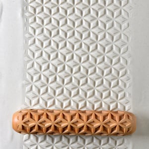 May include: A wooden rolling pin with a geometric pattern carved into the surface. The rolling pin is resting on a white surface with a similar geometric pattern.