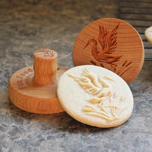 May include: Wooden cookie stamps with a bird design. One stamp is pressed into a cookie, showing the embossed image. The stamps are round and have a handle. The cookies are light-colored and sit on a dark surface.