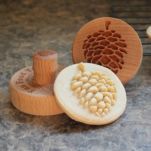 May include: Wooden cookie stamp set with a pine cone design. The set includes a round stamp with a handle, a round wooden mold with a pine cone carving, and a cookie with a pine cone imprint. The stamp is light brown.