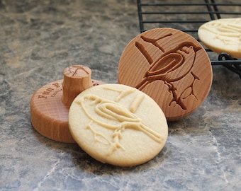 May include: Two wooden cookie cutters and a cookie with a bird design. The cookie cutters are round and have a bird on a branch design. The cookie is light brown and has a bird on a branch design.