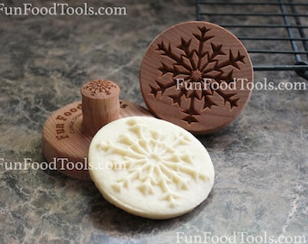 May include: Three wooden cookie stamps with snowflake designs. The stamps are used to create snowflake patterns on cookies. The stamps are made of wood and have a natural finish. The stamps are shown on a brown countertop.