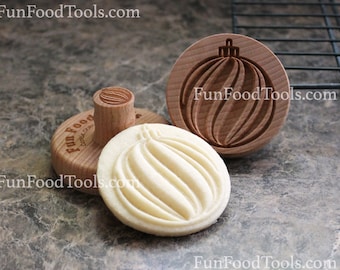 May include: Two wooden cookie stamps in the shape of a Christmas ornament. The stamps are made of light brown wood and have a detailed design. The smaller stamp is on top of a larger stamp. The smaller stamp is being used to imprint a design into a cookie. The cookie is white and has a swirl design. The text "FunFoodTools.com" is visible on the stamps.