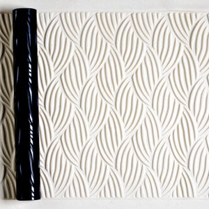 May include: A black rolling pin with a textured design. The rolling pin is on a white surface with a raised pattern of overlapping curved lines.