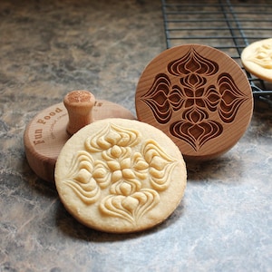 May include: A round wooden cookie stamp with a handle, featuring an intricate symmetrical floral pattern carved into the base. The stamp is positioned next to a light-colored cookie embossed with the same design and a wire cooling rack.
