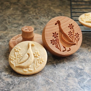 May include: A round wooden cookie stamp featuring a carved goose and flower design, accompanied by a light-colored baked cookie with the same embossed pattern. The stamp has a cylindrical handle and the text Wood Tools is visible on the base.