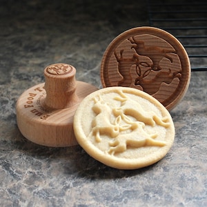 May include: A wooden cookie stamp with a handle and a round stamp, alongside a stamped cookie and a separate stamp. The stamp design features a rabbit motif. The cookie is light brown, and the stamps are made of light-coloured wood.