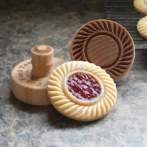 May include: A selection of wooden cookie stamps and a decorated biscuit with a red filling. The stamps are light brown and feature detailed designs. One stamp has a handle with the text "Food Tools". The biscuit has a fluted edge.