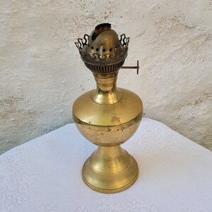 May include: A vintage brass oil lamp with a decorative, ornate top and a round base. The lamp is on a white tablecloth.