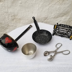 May include: A set of vintage kitchenware including a black and red ladle, a small black and white speckled frying pan, a silver measuring cup, a silver melon baller, and a black cast iron plaque with the text "GRACE BLESS O LORD THIS FOOD TO OUR USE AND MAKE US TO THY SERVICE AND MAKE US EVER MINDFUL OF THE NEEDS OF OTHERS IN HIS NAME. AMEN."