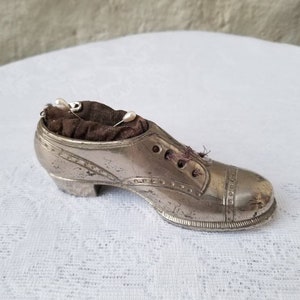 Pode incluir: Um almofadinha para alfinetes de prata em forma de sapato com um interior de tecido castanho. O almofadinha para alfinetes tem um design decorativo e um pequeno salto.