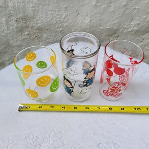 May include: Three glass drinking glasses with different designs. One glass has a yellow and green smiley face pattern, another has a red and white Peanuts character design, and the third has a black and white Peanuts character design.