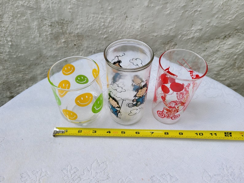 May include: Three glass drinking glasses with different designs. One glass has a yellow and green smiley face pattern, another has a red and white Peanuts character design, and the third has a black and white Peanuts character design.