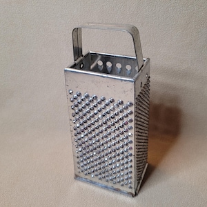 May include: A vintage, rectangular metal grater with a handle. The grater has multiple sides with different grating surfaces, including small, raised bumps and a fine grating area. The handle is a simple, arched design.