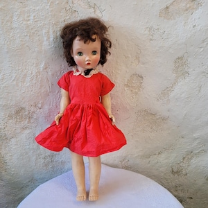 May include: A vintage doll with brown hair and a red dress. The doll has a white lace collar and is standing on a white surface.