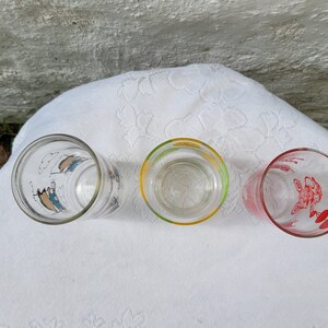 May include: Three vintage drinking glasses with colorful cartoon designs. The glasses are clear with a blue, yellow, and red rim. The designs are of a person in a boat, a person with a yellow umbrella, and a person with a red balloon.